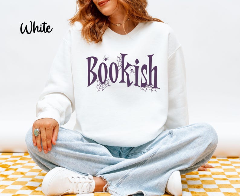 May include: A white long-sleeve crewneck sweatshirt featuring the word Bookish printed in a dark purple serif font. The design includes small black spider web illustrations and tiny spiders integrated into the lettering on the front chest.