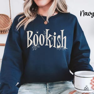 May include: A navy blue long-sleeve sweatshirt featuring the word "Bookish" printed in a cream-colored serif font. The design includes small spider web illustrations and spider graphics integrated around the letters. The text "Navy" appears in the top right corner.