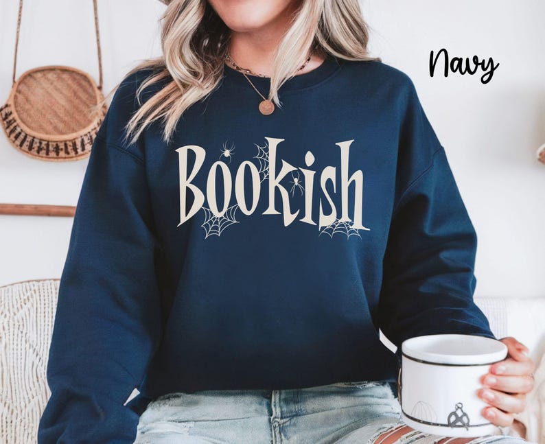May include: A navy blue long-sleeve sweatshirt featuring the word "Bookish" printed in a cream-colored serif font. The design includes small spider web illustrations and spider graphics integrated around the letters. The text "Navy" appears in the top right corner.