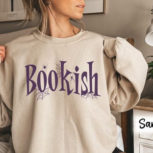 May include: A sand-colored crewneck sweatshirt featuring the purple text "BOOKISH" across the chest. The design includes purple spider web illustrations and small spider graphics integrated into the lettering. The sweatshirt has long sleeves.