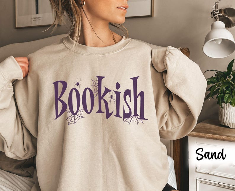 May include: A sand-colored crewneck sweatshirt featuring the purple text "BOOKISH" across the chest. The design includes purple spider web illustrations and small spider graphics integrated into the lettering. The sweatshirt has long sleeves.