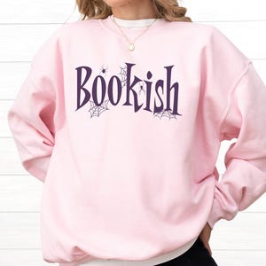 May include: A light pink crewneck sweatshirt featuring the word Bookish printed in dark purple lettering. The design includes small black spider web illustrations and tiny spider graphics integrated around the letters on the front of the garment.