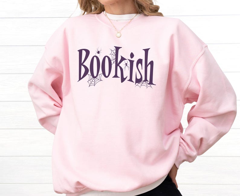 May include: A light pink crewneck sweatshirt featuring the word Bookish printed in dark purple lettering. The design includes small black spider web illustrations and tiny spider graphics integrated around the letters on the front of the garment.