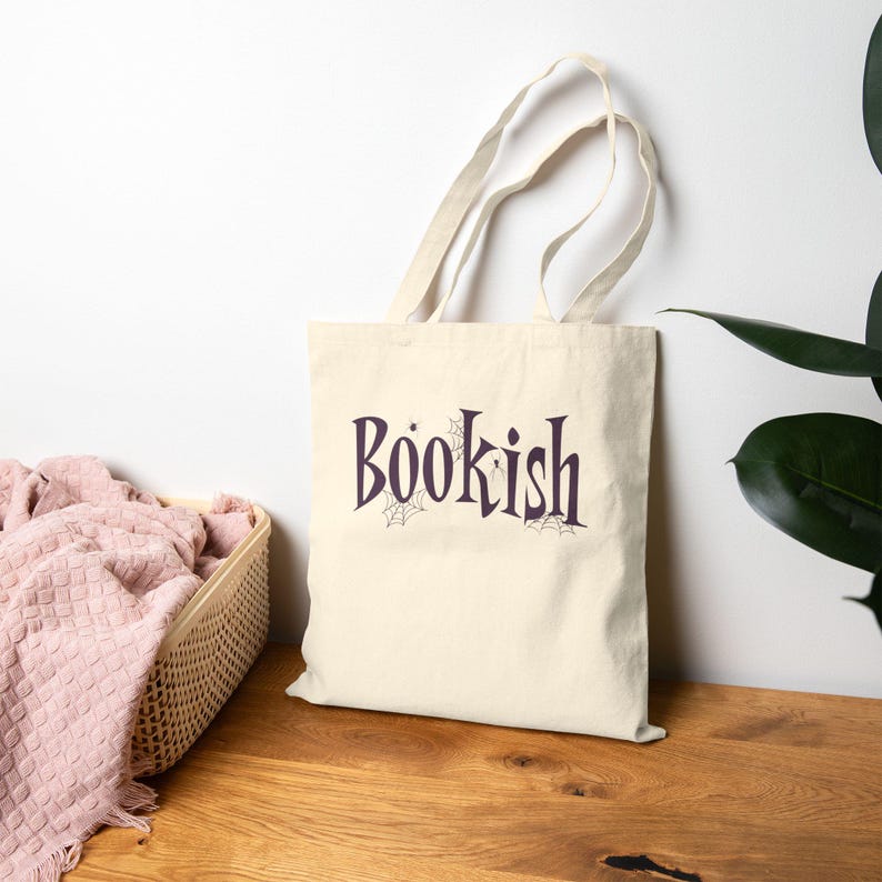 May include: A cream-colored canvas tote bag with two shoulder straps. The front features the word "BOOKISH" printed in a dark purple serif font, accented with small spiderweb illustrations and a tiny spider hanging from the letter B.