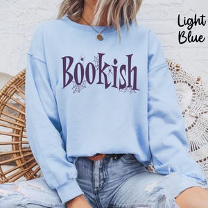 May include: A light blue crewneck sweatshirt featuring the dark purple text "BOOKISH" across the chest. The design includes small spider web and spider illustrations integrated into the lettering. The sweatshirt has long sleeves and a ribbed collar.