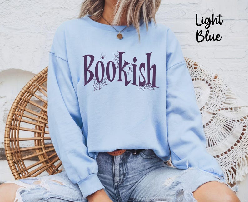 May include: A light blue crewneck sweatshirt featuring the dark purple text "BOOKISH" across the chest. The design includes small spider web and spider illustrations integrated into the lettering. The sweatshirt has long sleeves and a ribbed collar.