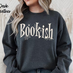 May include: A dark heather gray crewneck sweatshirt featuring the word "Bookish" printed in a large, cream-colored serif font. The design includes small, delicate spiderweb illustrations and tiny spiders integrated around the lettering.