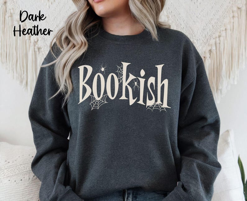 May include: A dark heather gray crewneck sweatshirt featuring the word "Bookish" printed in a large, cream-colored serif font. The design includes small, delicate spiderweb illustrations and tiny spiders integrated around the lettering.