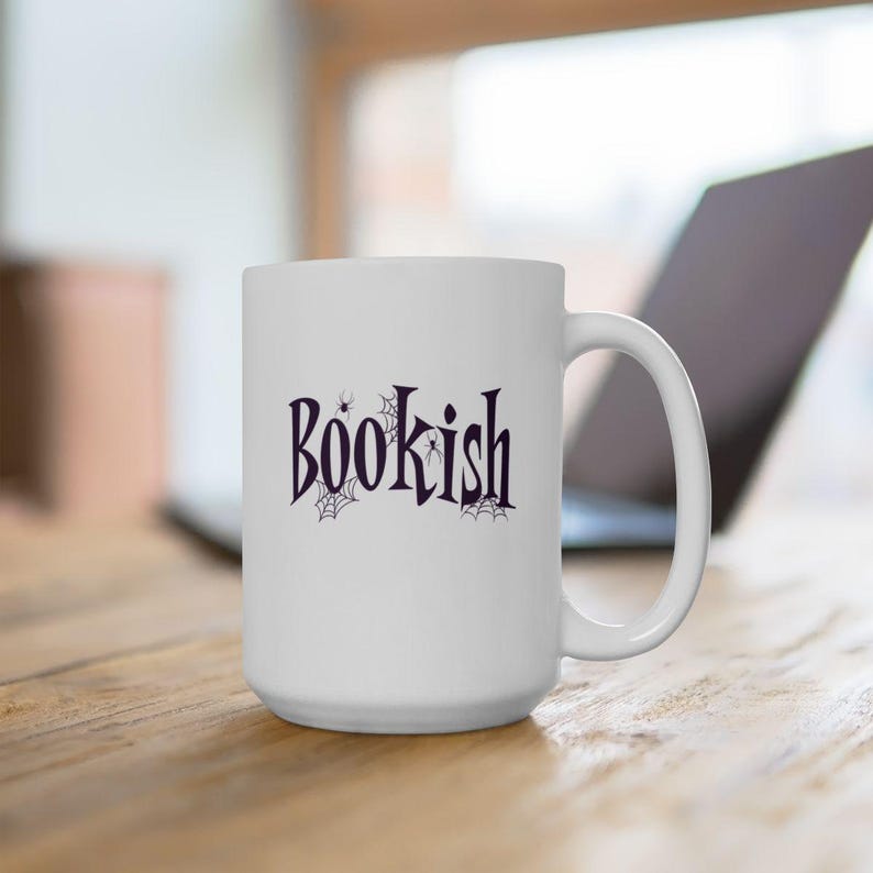 May include: A white ceramic mug featuring the word "BOOKISH" printed in a dark purple, stylized font. The text is decorated with small black spiderweb illustrations and tiny spiders integrated into the lettering on the front of the mug.
