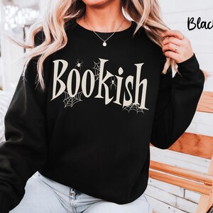 May include: A black long-sleeve sweatshirt featuring the word "Bookish" printed in a large, cream-colored serif font across the chest. The design includes small spider web illustrations and tiny spiders integrated into the lettering.