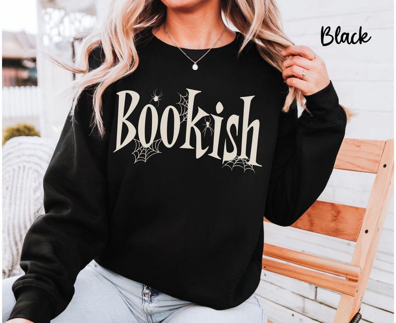 May include: A black long-sleeve sweatshirt featuring the word "Bookish" printed in a large, cream-colored serif font across the chest. The design includes small spider web illustrations and tiny spiders integrated into the lettering.