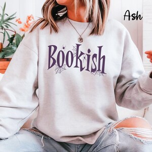 May include: An ash-colored crewneck sweatshirt featuring the word "Bookish" printed in dark purple lettering. The design includes small spider web illustrations and a spider integrated into the text. The sweatshirt has long sleeves and a ribbed collar.