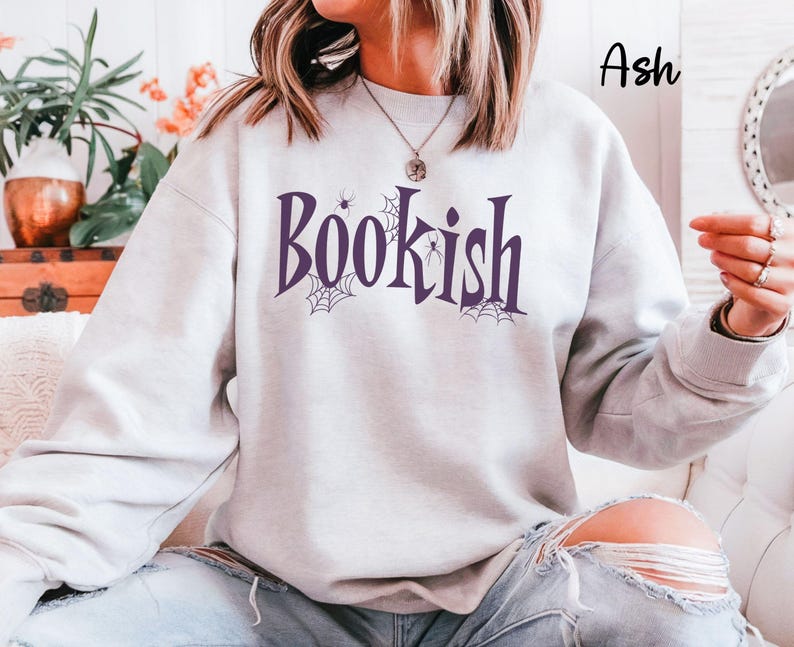 May include: An ash-colored crewneck sweatshirt featuring the word "Bookish" printed in dark purple lettering. The design includes small spider web illustrations and a spider integrated into the text. The sweatshirt has long sleeves and a ribbed collar.