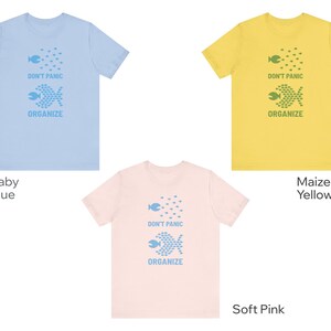 May include: Three t-shirts in baby blue, maize yellow, and soft pink. Each shirt features a graphic of fish and the words "DON'T PANIC ORGANIZE" in a matching color. The shirts are short-sleeved.