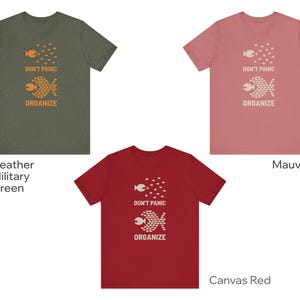 May include: Three t-shirts in heather military green, mauve, and canvas red. Each shirt features a graphic of two fish and the words "DON'T PANIC ORGANIZE" in orange or white. The shirts are short-sleeved.