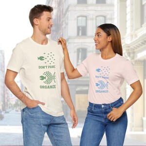 May include: Two people wearing matching t-shirts with the text "DON'T PANIC ORGANIZE" and a fish graphic. One shirt is cream-colored with green text and graphics, and the other is light pink with blue text and graphics. Both are wearing blue jeans.