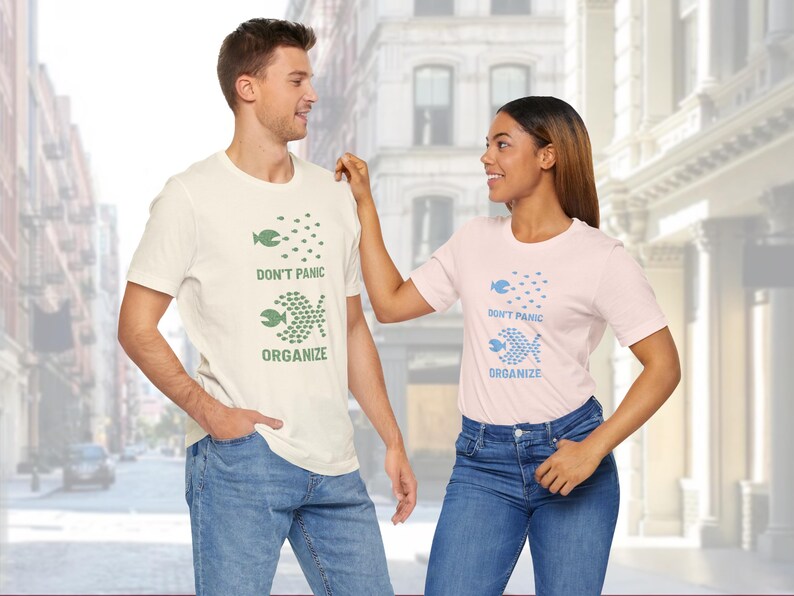 May include: Two people wearing matching t-shirts with the text "DON'T PANIC ORGANIZE" and a fish graphic. One shirt is cream-colored with green text and graphics, and the other is light pink with blue text and graphics. Both are wearing blue jeans.