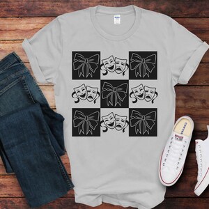 Könnte beinhalten: Ein graues T-Shirt mit einem schwarz-weißen Karomuster, das ein sich wiederholendes Design aus zwei Theatermasken und einer Schleife zeigt.
