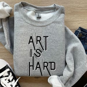 Op de afbeelding: Een grijze sweatshirt met de tekst "ART IS HARD" in een zwart-wit ontwerp. De sweatshirt is opgevouwen, met een spijkerbroek en een zwart-witte sneaker op de achtergrond.