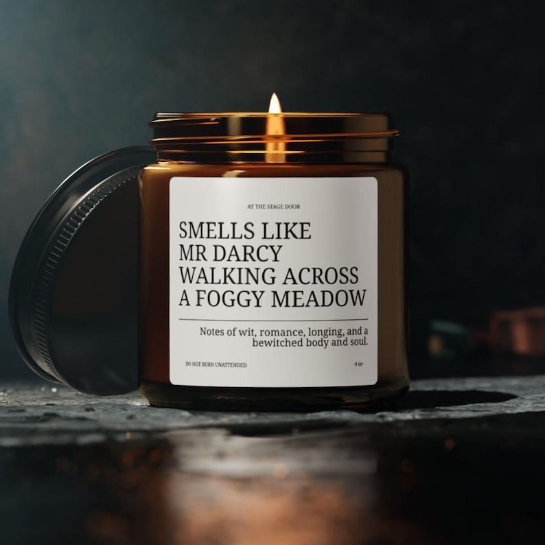 May include: A brown glass candle jar with a white label that reads "Smells Like Mr Darcy Walking Across A Foggy Meadow". The label also includes the text "Notes of wit, romance, longing, and a bewitched body and soul." and "Do Not Burn Unattended".