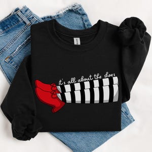 May include: Black sweatshirt featuring a graphic of red shoes and black and white striped legs. The text "it's all about the shoes" is printed on the sweatshirt.
