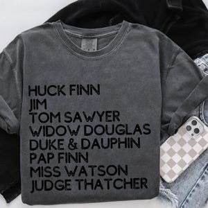 May include: A dark grey t-shirt with a list of characters from the novel The Adventures of Huckleberry Finn printed in black text. The characters listed are: Huck Finn, Jim, Tom Sawyer, Widow Douglas, Duke & Dauphin, Pap Finn, Miss Watson, and Judge Thatcher.
