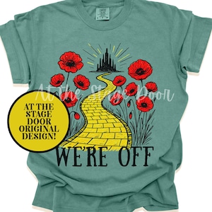 May include: Teal t-shirt with a yellow brick road design leading to a black castle, flanked by red poppies. The words "WE'RE OFF" are below the road. A yellow circle says "AT THE STAGE DOOR ORIGINAL DESIGN!"