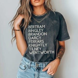 Puede incluir: Una camiseta gris oscura con un aspecto desgastado que presenta una lista de nombres en texto blanco: Bertram, Bingley, Brandon, Darcy, Ferrars, Knightly, Tilney, Wentworth.