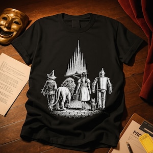 Op de afbeelding: Zwart t-shirt met een witte illustratie van personages uit The Wizard of Oz die naar een kasteel lopen. De illustratie toont de Vogelverschrikker, de Leeuw, Dorothy, de Blikken Man en Toto.
