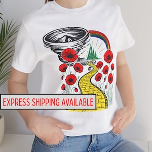May include: White t-shirt featuring a colorful graphic design inspired by The Wizard of Oz. The design includes a tornado, rainbow, yellow brick road, red poppies, and the Emerald City. The text "EXPRESS SHIPPING AVAILABLE" is displayed at the bottom.
