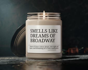 Peut inclure: Une bougie allumée dans un pot en verre avec un couvercle argenté. L'étiquette indique "SMELLS LIKE DREAMS OF BROADWAY" avec des notes de parfum. La bougie est sur une surface sombre avec un arrière-plan flou.