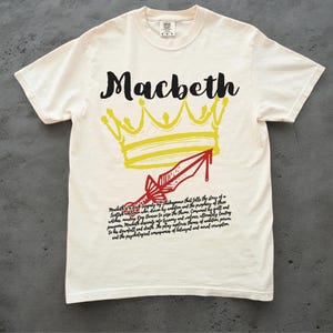 Peut inclure: T-shirt blanc avec une couronne jaune et une dague rouge avec une goutte de sang. Le texte "Macbeth" est écrit en noir au-dessus de la couronne. Le texte "Macbeth a dark tragedy by Shakespeare that tells the story of a Scottish general who is driven by ambition and the prophecy of three witches. Macbeth's ambition to seize the throne consumes him by guilt, fear, paranoia, murder, and descent into tyranny and violence. Ultimately leading to his downfall and death. The play explores themes of ambition, power, and the psychological consequences of betrayal and moral corruption." est écrit en noir sous la couronne.