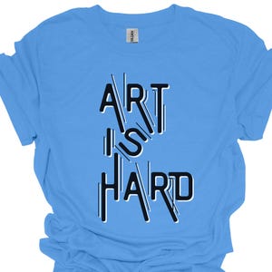 May include: A light blue t-shirt with the words "ART IS HARD" in a black, stylized font. The text has a white outline and a subtle shadow effect. The shirt is made of a soft material.