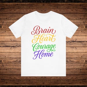 May include: White t-shirt with the words "Brain Heart Courage Home" in a rainbow of colors. The text is in a cursive font. The shirt is displayed on a wooden background.