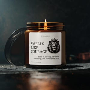 Puede incluir: Una vela encendida en un frasco de vidrio marrón con tapa dorada. La etiqueta dice "Smells Like Courage" con una ilustración de león. El texto "Notes of bravery, adventure, friendship, and happily ever after" también está en la etiqueta.