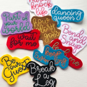 May include: A collection of colorful felt patches with embroidered text. Phrases include "Hard Knock Life," "Dancing Queen," "Part of Your World," and "Break a Leg." The patches are in various shapes and colors, with white stitching.