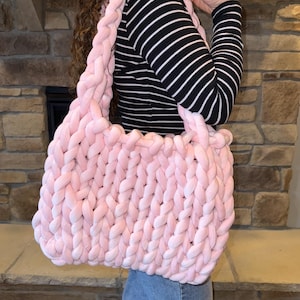 May include: A large, pink, chunky knit tote bag with a braided strap. The bag is made of soft, plush yarn and has a relaxed, casual style.