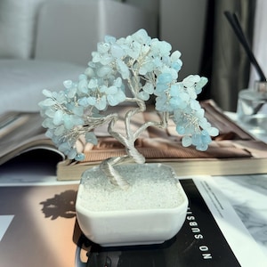 May include: A decorative bonsai tree sculpture with a silver wire trunk and branches. The tree's foliage is composed of small, light blue gemstones. The tree is planted in a white square pot filled with white pebbles, creating a unique home decor item.