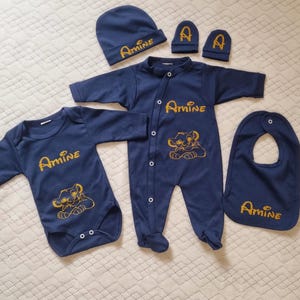 Peut inclure: Un ensemble de vêtements pour bébé bleu marine comprenant un body à manches longues, une grenouillère à boutons-pression, un bonnet, une paire de moufles et un bavoir. Chaque pièce porte le nom Amine et un motif de lionceau doré pailleté.
