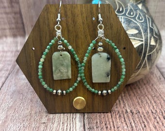 May include: A pair of handcrafted earrings displayed on a wooden stand. Each earring features a teardrop-shaped design with a central rectangular jade stone, surrounded by small green beads and silver accents. The earrings are suspended from silver hooks.