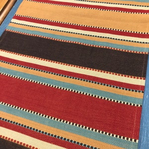 May include: Striped fabric with alternating bands of red, brown, beige, and blue. The fabric has a woven texture and a subtle pattern of small squares.
