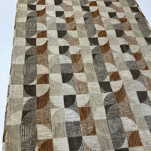 May include: Geometric patterned rug in shades of brown, beige, and gray. The rug features a repeating design of squares, semi-circles, and vertical lines. The rug is rectangular and appears to be made of woven material.