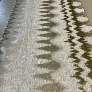May include: A long, rectangular table runner with a textured, woven design. The runner features a repeating pattern of olive green and gray geometric shapes against a light beige background. The edges have a zig-zag pattern.