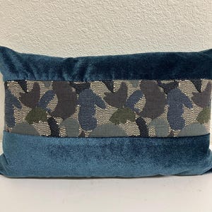 May include: Teal rectangular decorative pillow with a central band of patterned fabric. The band features a design of overlapping shapes in shades of blue, gray, and green, accented with silver beads. The pillow's main body is a solid teal velvet.