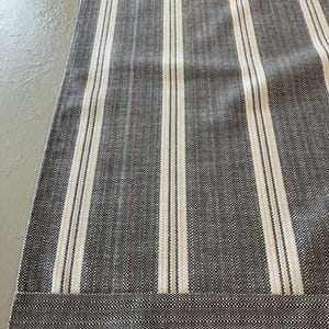 May include: A gray and white striped table runner. The runner features vertical white stripes against a gray background. The bottom edge has a horizontal stripe design. The fabric appears to be woven.