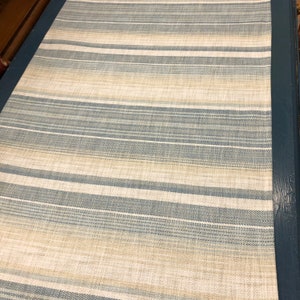 May include: A close-up of a striped fabric with alternating bands of beige and blue. The fabric has a subtle texture and a slightly distressed look.