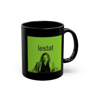 May include: Black coffee mug with a green square design featuring a portrait of a man with long hair and the word "lestat" in black text.