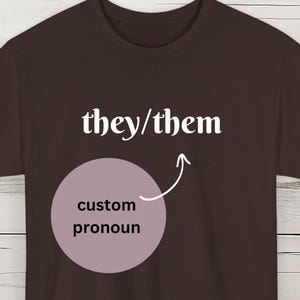 May include: Dark chocolate brown t-shirt with the text "they/them" above a light purple circle with the words "custom pronoun" and an arrow pointing to it. The words "DARK CHOCOLATE" are in a banner at the bottom.
