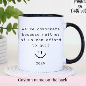 May include: A white ceramic mug with a black handle and rim. The front features black text that reads: we're coworkers because neither of us can afford to quit, a simple smiley face icon, and the year 2026 printed in a clean, sans-serif font.
