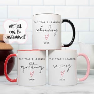 May include: Three white ceramic mugs with coloured handles and rims. The top mug has a black handle and reads THE YEAR I LEARNED embroidery 2026. The bottom left has a red handle and reads THE YEAR I LEARNED quilting 2026. The bottom right has a pink handle and reads THE YEAR I LEARNED sewing 2026.
